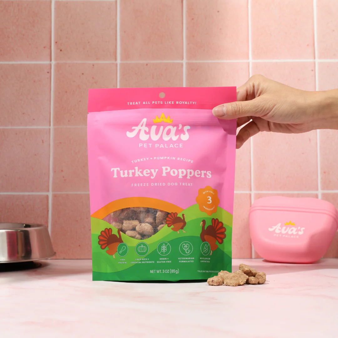 Ava's Pet Palace Turkey Poppers freeze dried turkey dog treats in pink and green packaging held by hand on kitchen counter