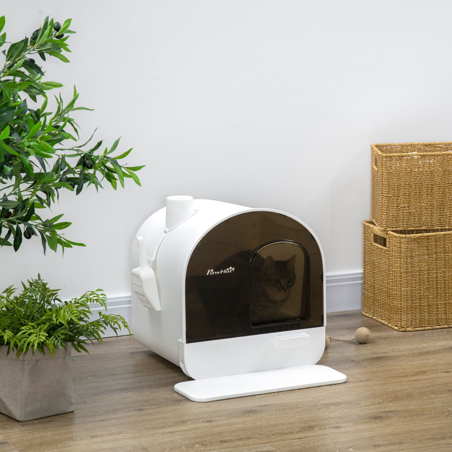 Covered cat litter box with lid in white and black with a cat inside placed on wooden floor near plants and wicker baskets